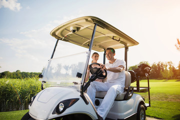 Happy man and woman are sitting in a white golf cart, which stands on the road of a golf club