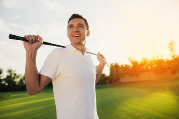A man in a white T-shirt is standing on a golf course and smiling, holding a golf club behind him