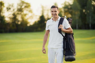 A man in a white suit walks around the golf course with a golf club bag and smiles