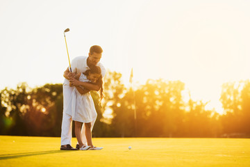 A man is teaching his daughter to play golf. He guides her, the girl is getting ready to make her first punch in golf