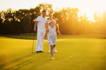 Happy family posing on a golf course on a sunset background. The girl smiles and runs towards the...