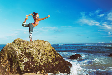 Meditation on sunset sky background. Young active woman stand in yoga pose on beach rock to keep...