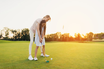 A woman is teaching a girl to play golf. The girl is preparing to hit, the woman is standing behind...