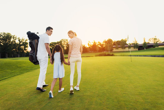 A man, a girl and a woman are walking along the golf course towards the sunset, holding hands