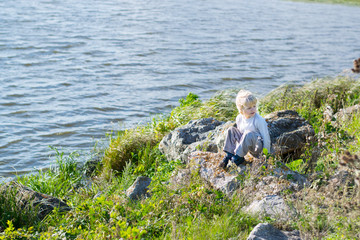 Fototapeta na wymiar Blond thoughtful boy sitting at a river bank on a rocks - Rusiian Urals wind sunny day