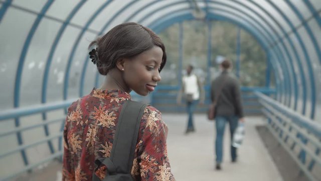 Young black female teacher with afro hairstyle wearing small leather jacket briefcase, walking out from office building, city streets, back view