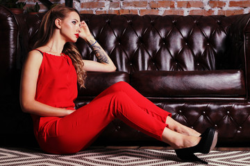 A sad girl is sitting next to the sofa, in a red dress and in heels