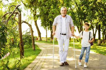 A boy and an old man on crutches are walking in the park. The boy is holding the old man's hand