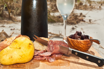 Chilled glass and bottle of vintage champagne, french bread, slices of salami, pear,  and a bowl of black olives on a bamboo serving plate on the sand of a tropical beach celebrating the autumn season