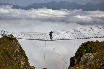 Man walking on suspension bridge and looking at cloudy mountains below.