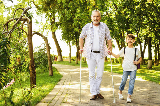 A boy and an old man on crutches are walking in the park. The boy is holding the old man's hand