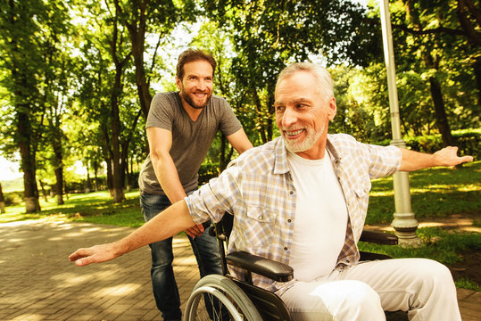 The old man on a wheelchair is walking in the park with his adult son. They fool around