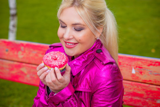 Blonde hair woman in a pink raincoat eats a donut and drinks coffee at park. Difficulties for diet. Cute girl on a diet struggles with a temptation to eat sweets