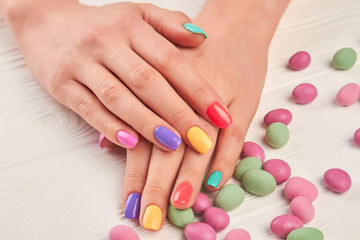 Obraz na płótnie Canvas Multicolored manicure and candies close up. Female hands with stylish colorful nails and multicolored candies on white wooden background close up.