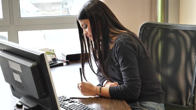 Stylish young businesswoman sitting at her desk in the office working on paperwork taking notes and making amendments