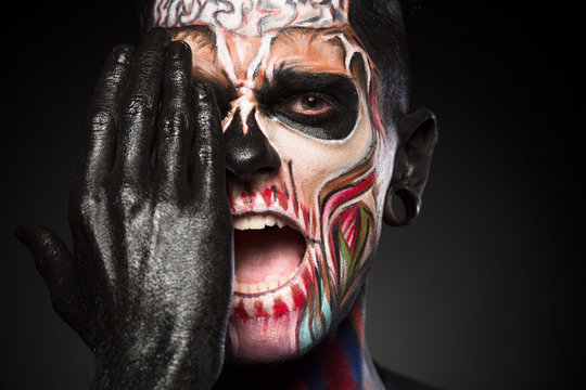 Halloween makeup concept, man covering zombie face with black painted hand. Detailed portrait of mans face with colores skeleton face. - Powered by Adobe