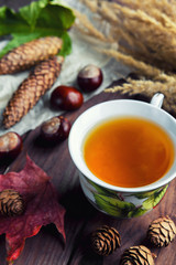 A cup with a hot tea drink on a wooden background with autumn leaves, cones, chestnuts
