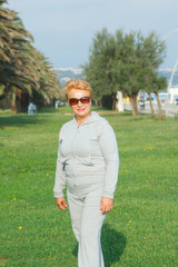 Adult woman on the background of the seaside resort. Senior woman in tracksuit enjoying life. Fitness and walking outdoors near the sea and the beach. Lifestyle adult retired. She is wear sunglasses.