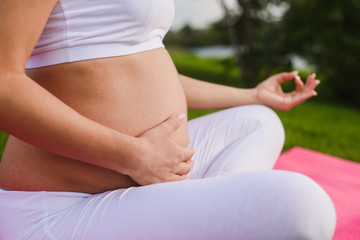 Fototapeta na wymiar Beautiful young pregnant woman doing yoga in park. Pregnant woman with hands over tummy. Close up.