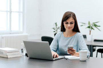 Young smiling business woman sitting at table in offce and using smartphone.