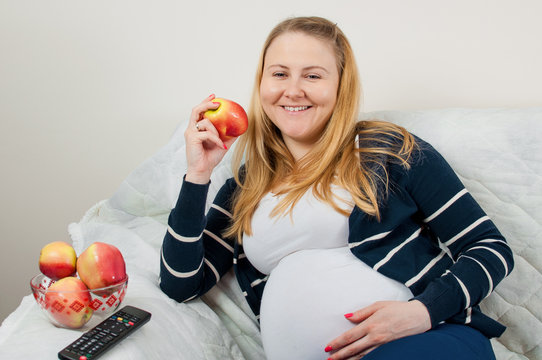 Beautiful pregnant woman watching TV on the sofa and eating fresh apples, horizontal image