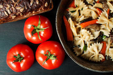 fresh cooked pasta with mushrooms and vegetables in a pan, along with tomatoes and bread with bran, horizontal image