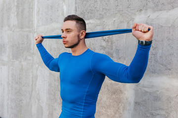 Muscular bodybuilder guy in a blue T-shirt doing exercises with sports gum on the background of gray wall. Fitness and healthy lifestyle concept