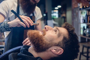 Side view headshot of a redhead bearded young man smiling ready for shaving in the hair salon, of a skilled barber with a classic straight razor in his hand