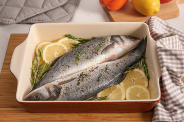 Baking tray with uncooked fish, lemon slices and herbs on kitchen table
