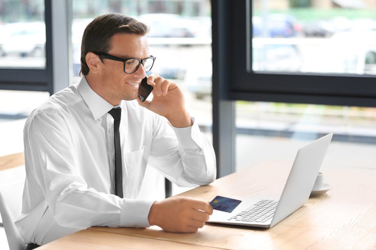 Young businessman holding credit card while making call at workplace
