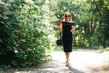 Young business lady in black dress and glasses with coffee and a bag walking in the park