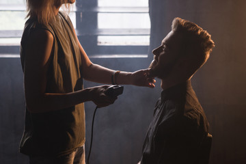 Female hairdresser shaving stylish man beard. Atmospheric barbershop dark background, smiling young...