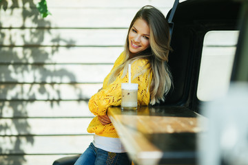 Pretty smiling woman is wearing yellow sweater drinking coffee latte near old retro bus