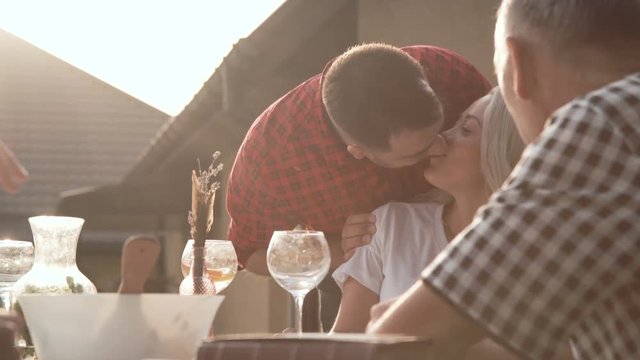 A beautiful woman tells something at the dinner table, meanwhile her husband comes up to her and kisses then tell something fun.
