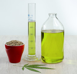 Hemp oil in a glass jar and beaker. Seeds of cannabis in a red cup and a leaf of marijuana.