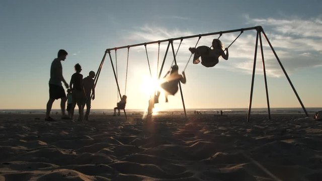 Silhouetted group of people enjoying a beautiful sunset at the beach on swing set.