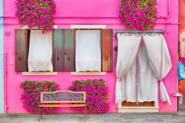 Fototapeta premium Różowy dom z różowymi kwiatami i roślinami. Ładna ławka pod oknami. Kolorowy dom w Burano wyspie blisko Wenecja, Włochy