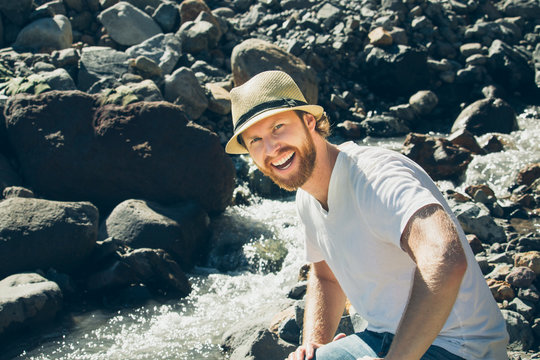 Bearded Blond Man Wearing Fedora And Smiling While Sitting On River Rocks