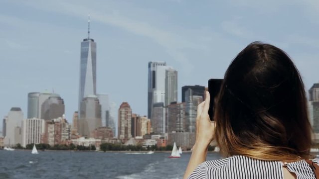 Young beautiful tourist girl taking photos of Manhattan in New York, America on smartphone from the boat on East river.