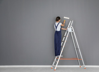 Young female decorator painting wall in room while standing on ladder - obrazy, fototapety, plakaty