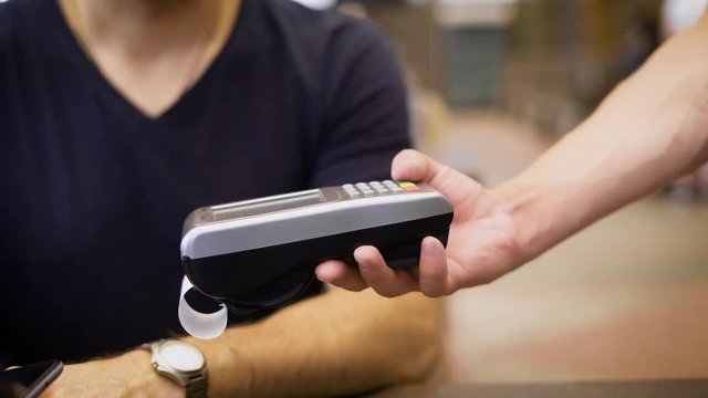 Close up of cashless payment in restaurant. Waiter giving to customer POS-terminal and cash voucher. Man in blue shirt making money transfer using his digital smartphone and NFC technology.