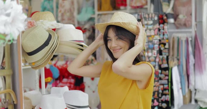 Young cheerful girl posing in shop of accessories trying on straw hat and looking at mirror while shopping in mall.