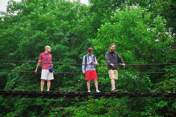 Young tourists stopped on suspension bridge across mountain river and photographed on a background of green trees.