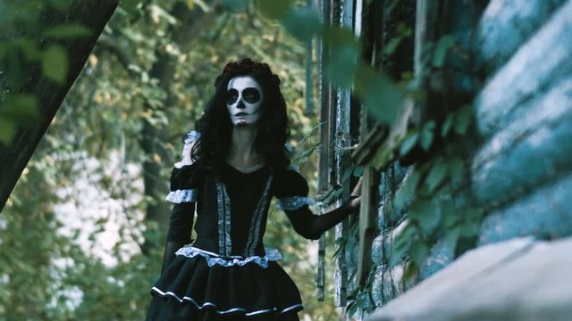 The Mexican Day of the Dead. The young woman with frightening make-up of skeleton for Halloween dressed in black clothes going along the wooden abandoned house. 4K