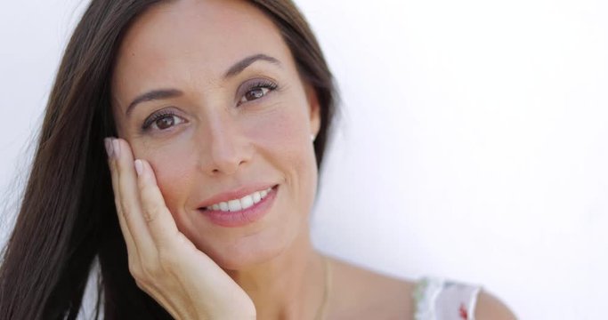 Portrait of young delightful model holding hand on face and looking gently at camera smiling slightly on white background.