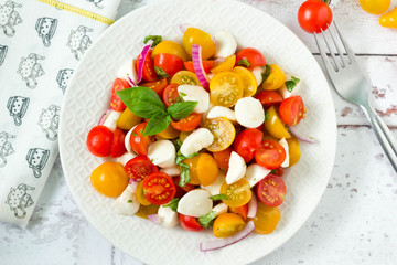 Homemade caprese salad with fresh yellow and red cherry tomatoes, mozzarella cheese, red onion and basil on kitchen table, top view.