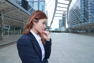 Wide angle shot of attractive young Asian business woman in suit thinking about her job at urban building background.