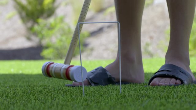 Detail of person using a mallet to hit a croquet ball through a hoop on a bright green lawn in summer. Slow motion 4K recorded at 60fps.