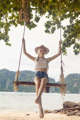 Gorgeous young woman is swinging on a swing which hangs on a tree and enjoying the beautiful summer weather on the island with a beautiful view.