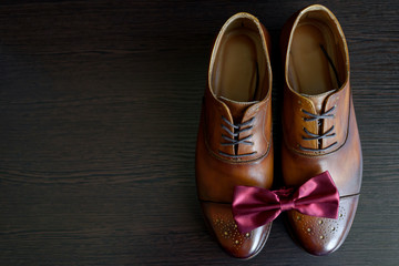 Close up of burgundy bowtie on brown leather shoes, free space. Modern man or groom accessories. Wedding details. Men's casual outfits with shoes and bowtie on wood background